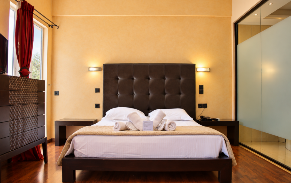 Anastazia Luxury Suites spacious family room featuring a dark tufted headboard king bed with clean white linens and a modern glass shower layout