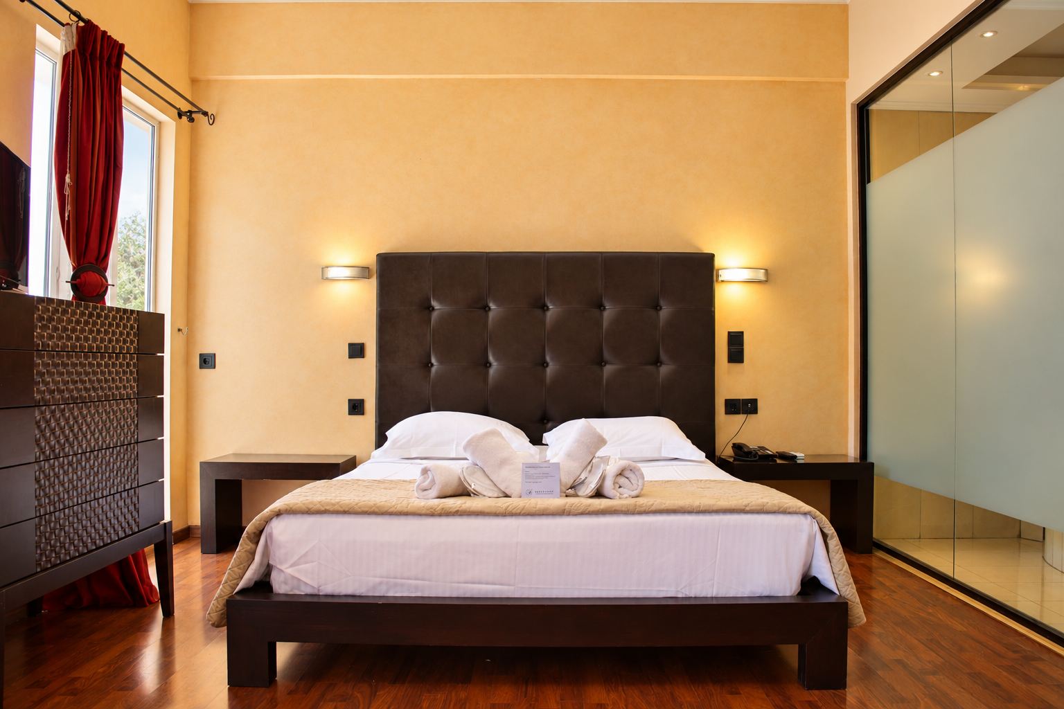 Anastazia Luxury Suites spacious family room featuring a dark tufted headboard king bed with clean white linens and a modern glass shower layout