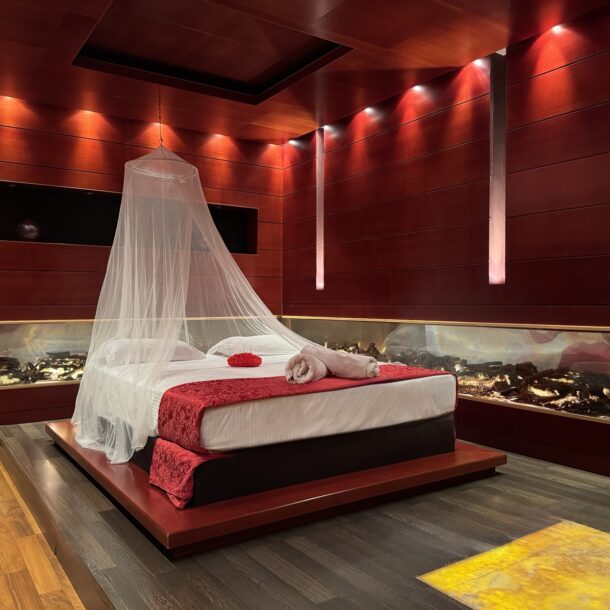 Anastazia Luxury Suites Deluxe Suite featuring a romantic canopy platform bed, red ambient lighting, mahogany wood panels, and a built-in panoramic fireplace in Drosia