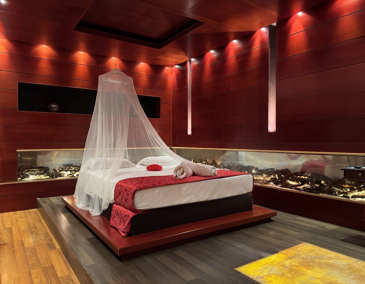 Anastazia Luxury Suites Deluxe Suite featuring a romantic canopy platform bed, red ambient lighting, mahogany wood panels, and a built-in panoramic fireplace in Drosia