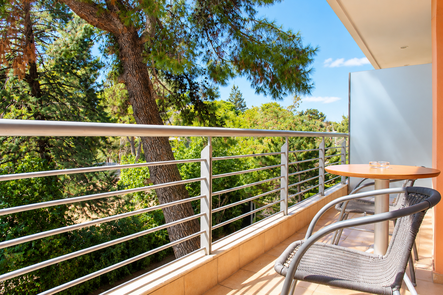 Sunlit private balcony of the Family Room Suite at Anastazia Luxury Suites & Spa in Drosia, featuring modern metal railings, outdoor seating furniture, and a relaxing, scenic view of the surrounding lush pine trees.