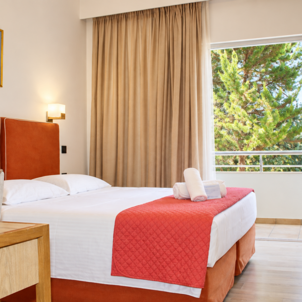 Anastazia Luxury Suites brightly lit Comfort Room layout featuring a bed with a red headboard, clean white linens, wood furniture, and a sunlit balcony window view in Drosia