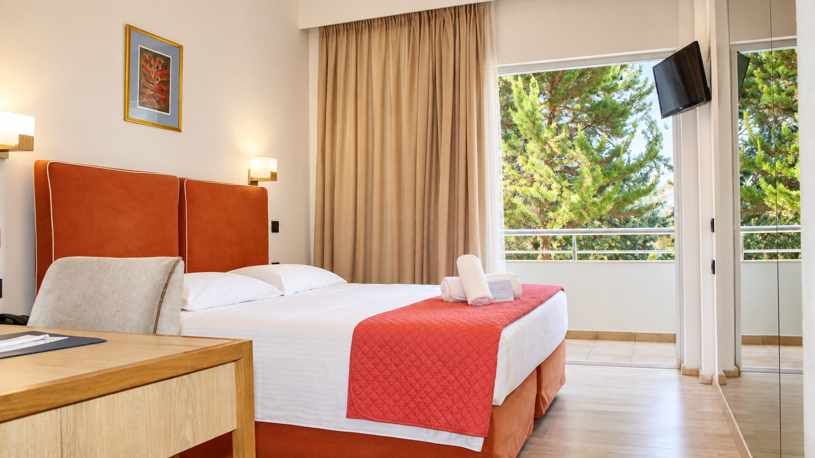 Anastazia Luxury Suites brightly lit Comfort Room layout featuring a bed with a red headboard, clean white linens, wood furniture, and a sunlit balcony window view in Drosia